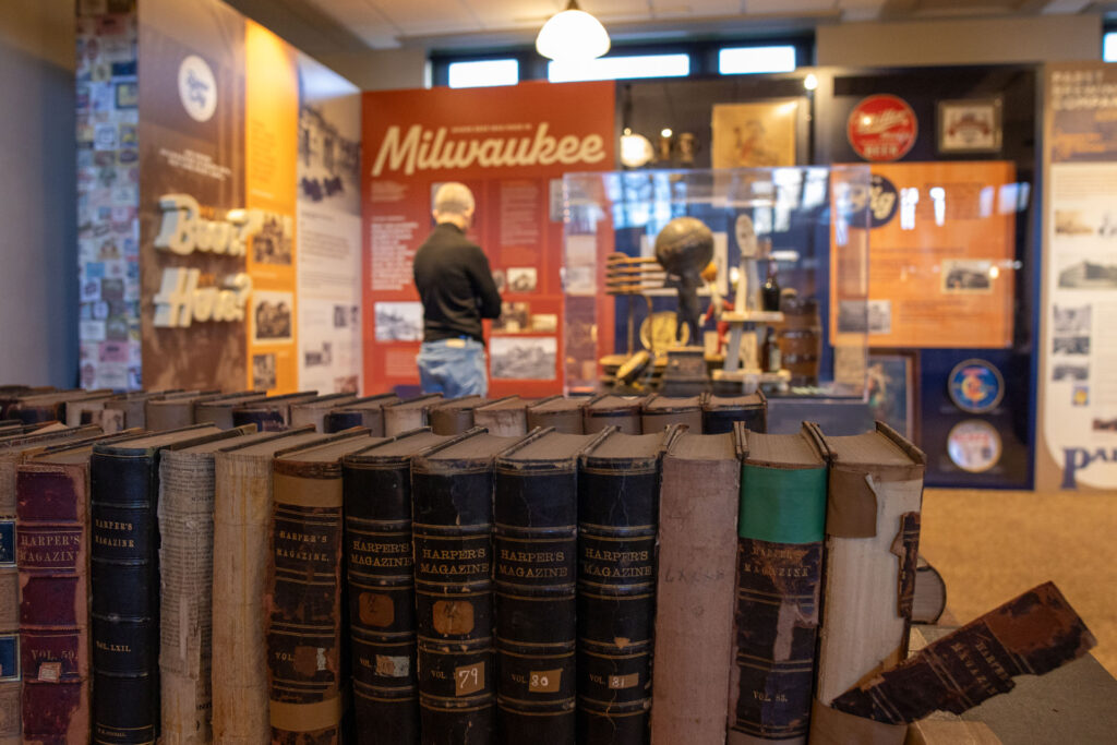 Rows of historic Harper’s Magazine volumes with a visitor reading Milwaukee history exhibits behind them.
