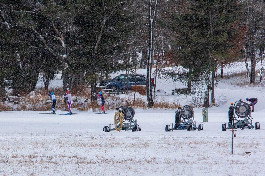 Cross-country skiers move across snowy trails at Lapham Peak while snowmaking machines sit in the foreground during snowfall.
