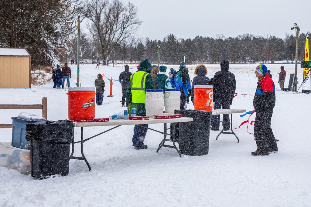 Volunteers stand near a refreshment table with sports drink coolers during snowfall at the Lapham Loppet.